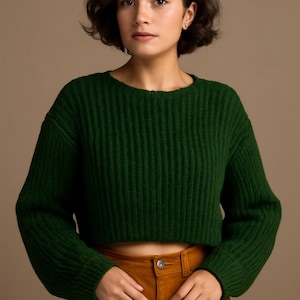 Puede incluir: Un suéter corto verde bosque con una textura de punto acanalado. El suéter tiene mangas largas y cuello redondo. La modelo lleva el suéter con pantalones de cintura alta color óxido.