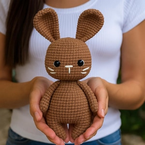 Puede incluir: Un conejito de ganchillo marrón hecho a mano con ojos negros y bigotes blancos. El conejito tiene orejas largas y se sostiene con las manos abiertas. El juguete está hecho de un hilo texturizado y tiene un diseño suave y afelpado.