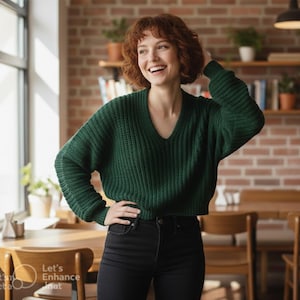Puede incluir: Una mujer con un suéter verde oscuro con cuello en V y jeans negros sonríe en un café. El suéter tiene una textura de punto acanalado. El fondo incluye una pared de ladrillos, mesas de madera y una ventana.