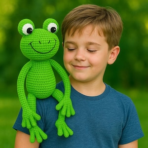 Op de afbeelding: Een felgroene gehaakte kikkerpop met grote ogen en een lachende mond. De kikker rust op de schouder van een persoon. De armen en benen van de kikker zijn uitgestrekt. De persoon draagt een blauw t-shirt.