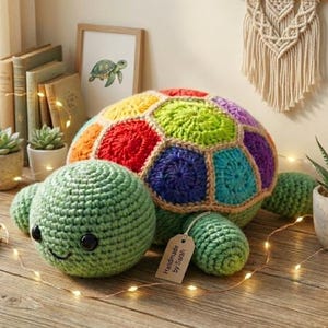 Puede incluir: Una tortuga de ganchillo hecha a mano, colorida, con cabeza y patas verdes, y un caparazón multicolor. El caparazón presenta formas geométricas rojas, naranjas, amarillas, verdes, azules y moradas. La etiqueta dice "Handmade by Sarah".