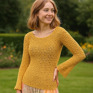 May include: Mustard yellow crochet top with a scoop neck and long sleeves. The top has a textured diamond pattern and flared sleeves. The model is wearing a floral patterned skirt. The background is a blurred green garden.
