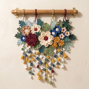 Puede incluir: Un tapiz de pared que presenta una colección de flores y hojas de ganchillo en tonos crema, burdeos, azul, amarillo y verde. El arreglo está suspendido de una barra de madera horizontal con soportes de latón y cuatro cordones trenzados.