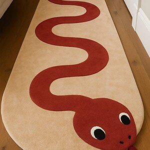 May include: A beige runner rug with a red snake design. The snake's head is at one end, featuring black eyes and a red tongue. The rug is placed on a wooden floor.