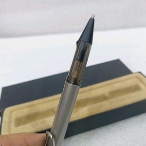 以下が含まれることがあります： メタリックシルバーのバレルと半透明のダークグレーのグリップセクションを備えたボールペン。マットブラックの円錐形のペン先と、バレルの側面に固定されたシルバーカラーの金属製クリップが付いており、黒い収納ケースと一緒に提示されています。
