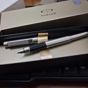 Puede incluir: Una pluma estilogr&aacute;fica Parker plateada con un agarre negro y un capuch&oacute;n plateado a juego, presentada en una caja beige con el logotipo de Parker. La pluma se muestra sobre una superficie negra, junto a un teclado.