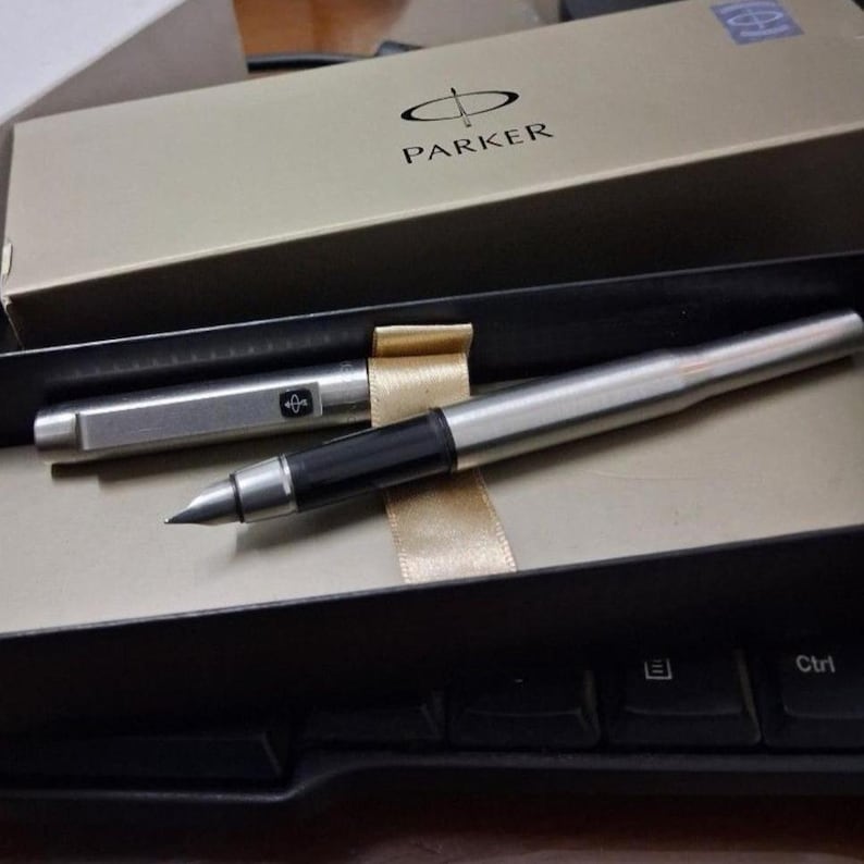 Puede incluir: Una pluma estilogr&aacute;fica Parker plateada con un agarre negro y un capuch&oacute;n plateado a juego, presentada en una caja beige con el logotipo de Parker. La pluma se muestra sobre una superficie negra, junto a un teclado.