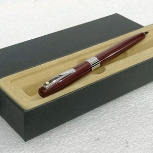 Può includere: Una penna bordeaux con dettagli argentati è esposta in una scatola nera con interno beige. La penna ha un design elegante, con una fascia e una clip argentate. La penna è presentata in una scatola rettangolare, adatta per essere regalata o come articolo premium.
