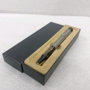 以下が含まれることがあります： 黒いグリップと銀色のポケットクリップが付いたメタリックシルバーのボールペン。ベージュの成形インサートが入った長方形の黒いギフトボックスに収められています。ペン先付近には暗い半透明のセクションがあり、黒いエンドキャップが付いています。
