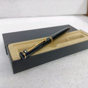 Op de afbeelding: Een zwarte vulpen met goudkleurige accenten, waaronder een clip en sierlijsten, ligt in een zwarte presentatiedoos met een beige interieur. De pen heeft een slanke, gepolijste uitstraling, wat duidt op een luxe schrijfinstrument.