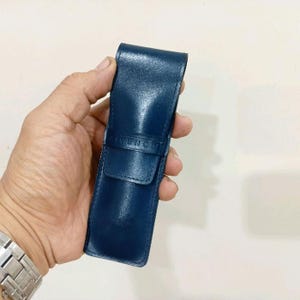 May include: A navy blue leather pen case with a flap closure. The case has the brand name "GIVENCHY" embossed on the front. The case is held in a person's hand.