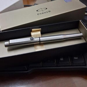 Puede incluir: Un bol&iacute;grafo Parker plateado en su caja original. El bol&iacute;grafo es cil&iacute;ndrico con un clip rectangular y una banda negra cerca de la punta. La caja es beige con el logotipo de Parker en negro.