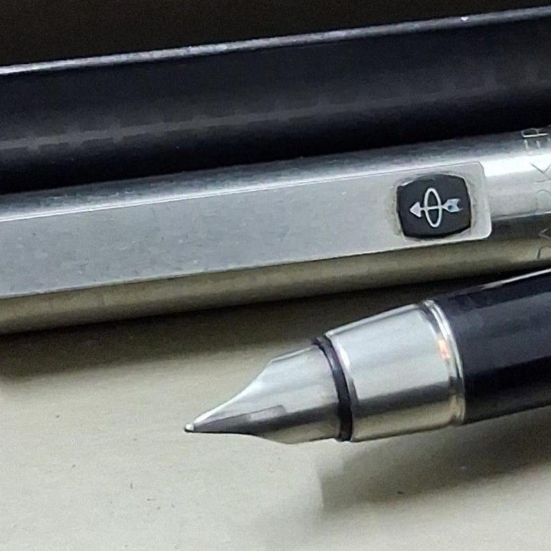 Puede incluir: Primer plano de una pluma estilogr&aacute;fica plateada y negra. El bol&iacute;grafo tiene un cuerpo plateado con un capuch&oacute;n negro y un plum&iacute;n plateado. El bol&iacute;grafo presenta un peque&ntilde;o emblema con un dise&ntilde;o de flecha. Es un instrumento de escritura.