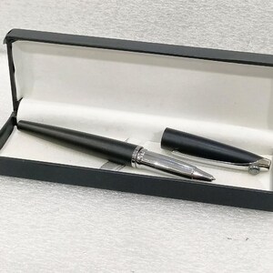 Pode incluir: Uma caneta preta com ponta e detalhes prateados, apresentada numa caixa preta aberta com interior branco. A tampa da caneta está separada. Um instrumento de escrita elegante e profissional.