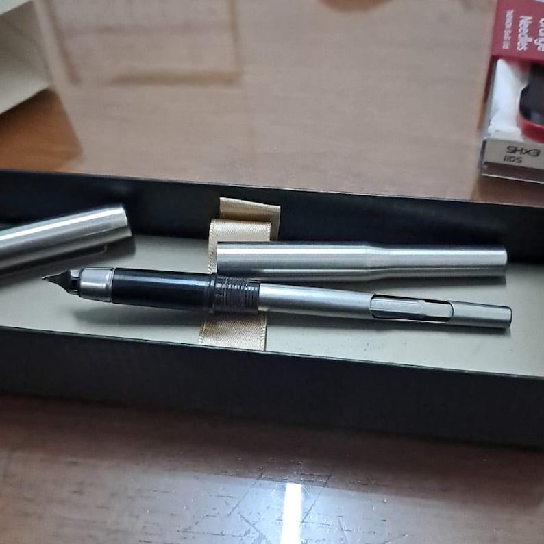 Puede incluir: Una pluma estilogr&aacute;fica plateada con agarre y plum&iacute;n negros, presentada en una caja negra. La pluma est&aacute; parcialmente desmontada, con el capuch&oacute;n y el cuerpo separados. La caja tiene una cinta de raso.
