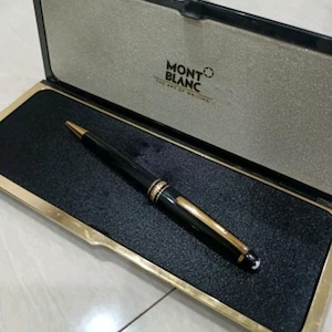 May include: A black Montblanc pen with gold-colored accents rests inside its open case. The case is black with a gold border and a gray interior. The Montblanc logo and text are visible on the case lid.