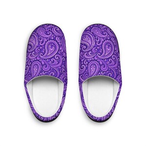 May include: A pair of purple paisley pattern slippers. The slippers have a white interior and a black trim. The paisley design features swirling floral elements in shades of purple, creating a decorative and comfortable footwear option.