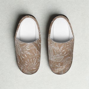 Puede incluir: Un par de zapatillas beige con un estampado floral blanco. Las zapatillas tienen un interior blanco y un ribete negro. El diseño floral incluye hojas y otros elementos botánicos.