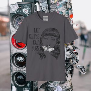 May include: Charcoal grey t-shirt with a black and white graphic of a person with a butterfly. The text "LET ELITES EAT BUGS" is printed on the front. The t-shirt has a crew neck and short sleeves.