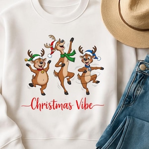 May include: White sweatshirt featuring a festive design with three dancing reindeer wearing Christmas hats and scarves. The phrase "Christmas Vibe" is written in red script below the reindeer. A fun, holiday-themed apparel item.