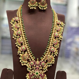 May include: An ornate gold necklace and earring set with a detailed design. The necklace has a central pendant and repeating floral patterns with red and green gemstones. The earrings match the necklace. The jewellery is displayed on a brown mannequin.
