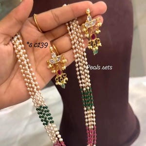 Puede incluir: Un conjunto de collar y pendientes de perlas con detalles dorados. El collar presenta hilos de perlas blancas con secciones de cuentas verdes y rosas. Los pendientes tienen piedras claras y rojas. El texto "Peals sets" es visible.