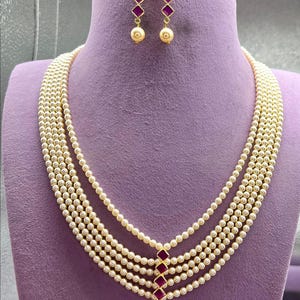Può includere: Un set di collana e orecchini di perle esposto su un manichino viola. La collana presenta più fili di piccole perle rotonde, con un ciondolo con accenti rossi e dorati. Gli orecchini hanno un design abbinato.