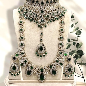 May include: An elaborate jewellery set comprising a necklace, earrings, and a headpiece. The set is embellished with emerald green gemstones and clear crystals, set in a silver-toned metal. The necklace and earrings have a drop design, and the headpiece has a central pendant.
