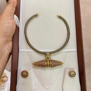 Puede incluir: Un collar dorado con un cordón negro envuelto en espiral, centrado con un colgante bicónico incrustado con pequeñas piedras rojas en un diseño de celosía. El conjunto incluye dos pendientes circulares a juego con piedras rojas.