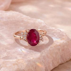 Könnte beinhalten: Ein goldener Ring mit einem großen ovalen Rubin als Mittelpunkt. Der Ring hat kleinere Akzentsteine auf beiden Seiten des Rubins. Er liegt auf einer hellrosa Steinoberfläche mit einem weichen, diffusen Hintergrund.