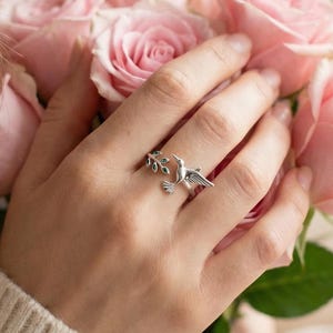 Könnte beinhalten: Ein silberner Ring mit einem Vogelmotiv, einschließlich ausgebreiteter Flügel und einem kleinen Zweig. Der Ring befindet sich an einem Finger, der einen Strauß rosa Rosen hält. Die Person trägt einen cremefarbenen Pullover.