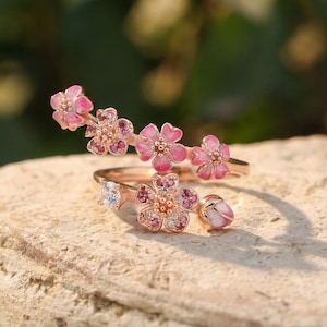 Könnte beinhalten: Zwei roségoldene Ringe mit rosa Blumendesigns. Ein Ring hat eine Reihe kleiner rosa Blüten, der andere eine größere Blüte mit einem klaren Stein und rosa Akzenten. Die Ringe liegen auf einer strukturierten Steinoberfläche.