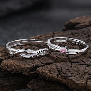 Op de afbeelding: Twee verstelbare zilveren ringen. Eén ring heeft een roze edelsteen, de andere heeft drie kleine, heldere edelstenen. Beide ringen hebben een delicaat, verweven ontwerp. De ringen worden gepresenteerd op een stuk donker, getextureerd hout.