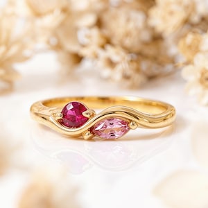 May include: A gold ring with a round, deep pink gemstone and a marquise-cut, light pink gemstone. The ring has a flowing design with delicate details. The background is soft and blurred.