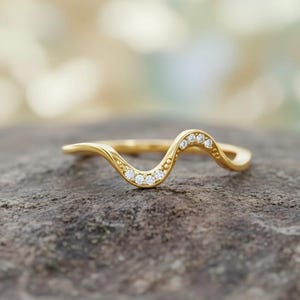 Könnte beinhalten: Ein goldfarbener Ring mit wellenförmigem Design. Der Ring hat eine Reihe kleiner, klarer Edelsteine, die entlang der oberen Kurve gesetzt sind. Der Ring wird auf einer strukturierten, grauen Oberfläche mit unscharfem Hintergrund gezeigt.
