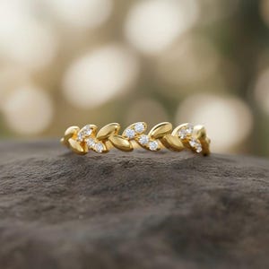 Puede incluir: Un anillo dorado con diseño de hojas, cada una adornada con pequeñas gemas transparentes. El anillo se encuentra sobre una superficie oscura y texturizada, con un fondo borroso y claro.