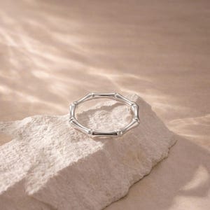May include: A silver ring featuring a bamboo design. The ring is crafted from metal and comprises a series of connected bamboo-shaped segments. It's presented on a white, textured surface, set against a neutral backdrop.