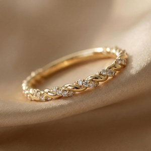 May include: A gold-colored ring with a braided design, adorned with small, clear gemstones. The ring is set against a soft, neutral-toned background, highlighting its intricate details and elegant appearance. The ring's band is approximately 0.1 inches wide.