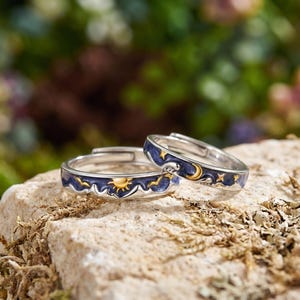 May include: Two silver rings with celestial designs. One ring has a gold sun, the other a gold moon and stars. Both have a dark blue enamel band with gold accents. The rings are displayed on a natural stone surface.