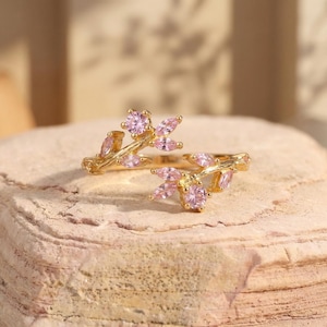 Pode incluir: Um anel dourado com um design floral. O anel apresenta detalhes de pedras preciosas rosa em um padrão semelhante a um galho. As pedras preciosas são redondas e em forma de marquesa, cravejadas em uma faixa texturizada.