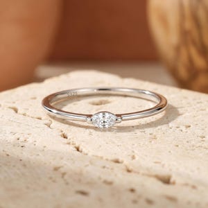 May include: A slender silver ring featuring a marquise-shaped diamond. The ring band is thin and polished, with the hallmark "S925" visible. The diamond is set horizontally, enhancing the ring's minimalist and elegant design.