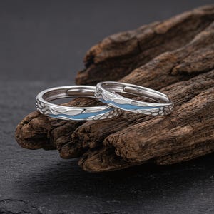 May include: Two silver rings with a wave design and a light blue enamel inlay. The rings are displayed on a piece of dark, textured wood. The rings are likely adjustable.