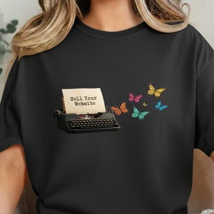 May include: Black t-shirt featuring a vintage typewriter with the text "Sell Your Website" printed on a paper. Colorful butterflies are flying away from the typewriter. The shirt is a dark color.