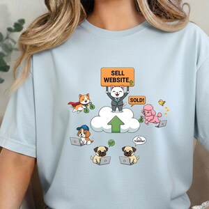 May include: Light blue t-shirt featuring a cartoon design of dogs working on laptops. A cat holds a sign that says "SELL WEBSITE" and "SOLD!". Other dogs are shown with money and a cloud with an upward arrow. A cute duck is also shown.