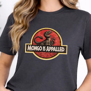 May include: Dark gray t-shirt featuring a dinosaur graphic inside a circular logo with the text "MONGO IS APPALLED". The dinosaur is brown and the logo has red and yellow accents. The shirt is a short-sleeved crew neck.
