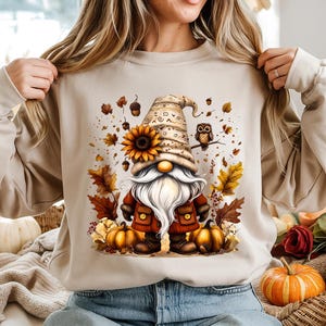 Peut inclure: Sweat-shirt beige avec un motif de gnome fantaisiste. Le gnome a une longue barbe blanche, un tournesol et un chapeau pointu. Des feuilles d'automne, des citrouilles et un hibou complètent le thème automnal. Un haut confortable et festif.
