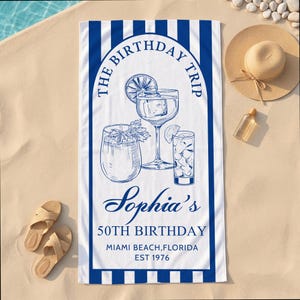 Könnte beinhalten: Weißes Strandtuch mit blauen Streifen und einer blauen Illustration von Cocktails. Der Text enthält "The Birthday Trip", "Sophia's 50th Birthday", "Miami Beach, Florida, EST 1976". Ein Strohhut, Sandalen und eine Flasche liegen auf dem Sand.
