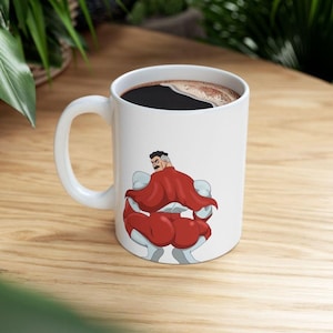 May include: White ceramic coffee mug with a handle, filled with dark liquid. The mug features a cartoon illustration of a superhero in a red cape and white and gray suit. The mug is on a wooden surface.