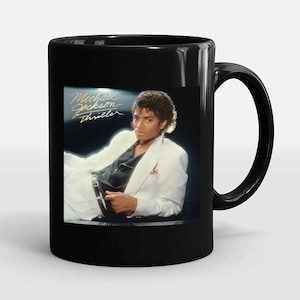May include: Black ceramic mug featuring a portrait of Michael Jackson from the Thriller album cover. The image shows Jackson in a white suit and the album title in gold script. A classic music-themed gift.