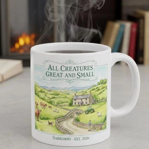 May include: White ceramic mug with a handle, featuring a countryside illustration with the text "All Creatures Great and Small" and "Darrowby - Est. 2026". The mug contains a dark liquid, with steam rising from it. The illustration depicts a farm scene.